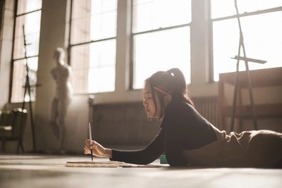 Young woman painting while lying on the floor in a sunlit art studio.