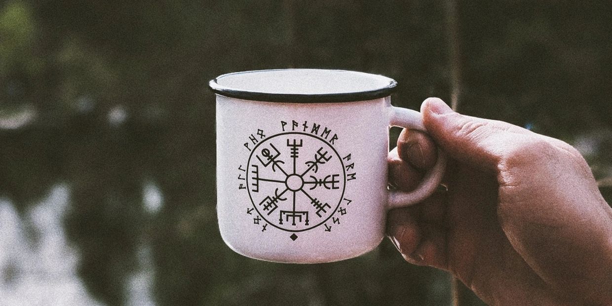 Hand holding a white mug with Viking runes and symbols.