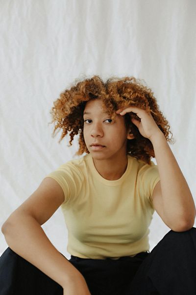 A youth with long, large curly afro  - light brown, gold in color faces the camera. Hand on head.