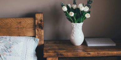 White tulips in a vase on a wooden bedside table next to a bed.