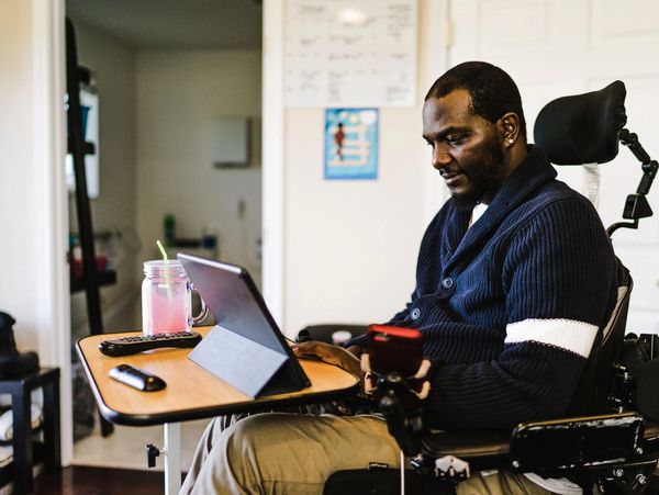 Man in wheelchair using tablet at a desk with a drink nearby.
