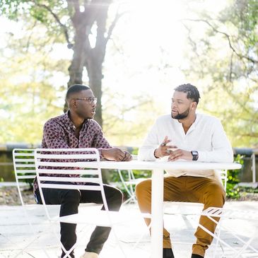 Two black men have a discussion outdoors on a white table. This is a coaching conversation 