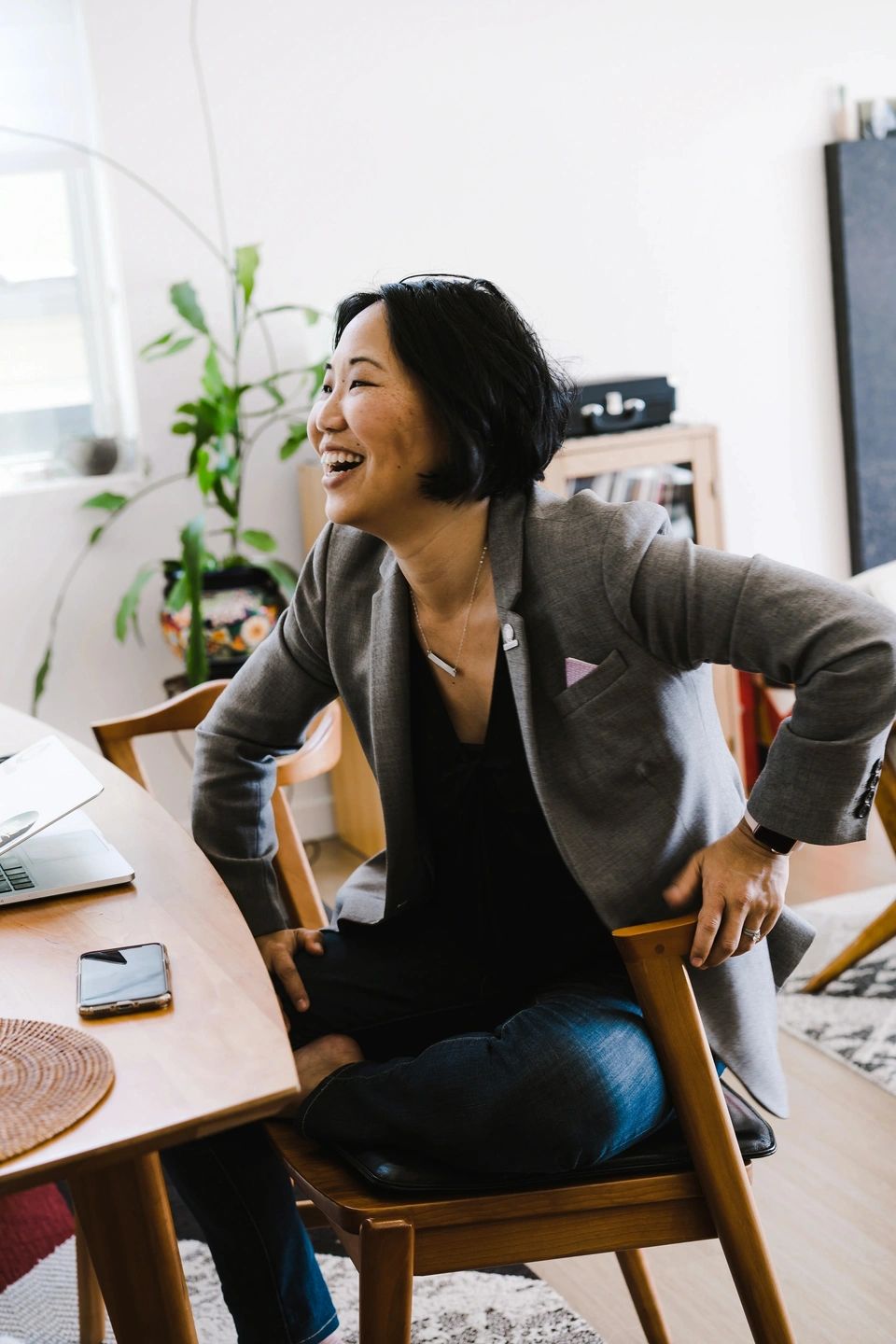 Woman smiling while seated at a wooden table, engaged in conversation, with a laptop and smartphone nearby, in a bright, modern workspace.