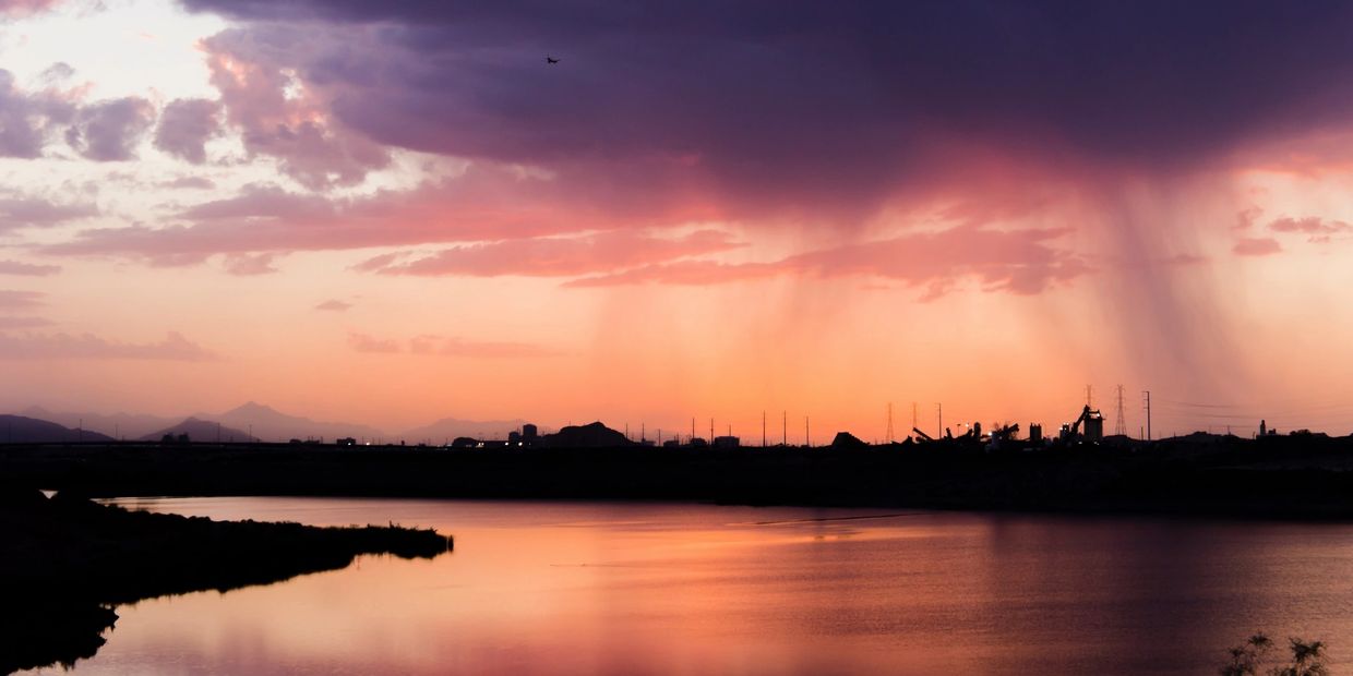 Sunset with rain showers over a calm body of water and silhouetted industrial structures.
