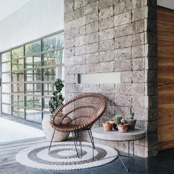 Cozy modern corner with a rattan chair, round rug, and potted plants beside a stone wall.