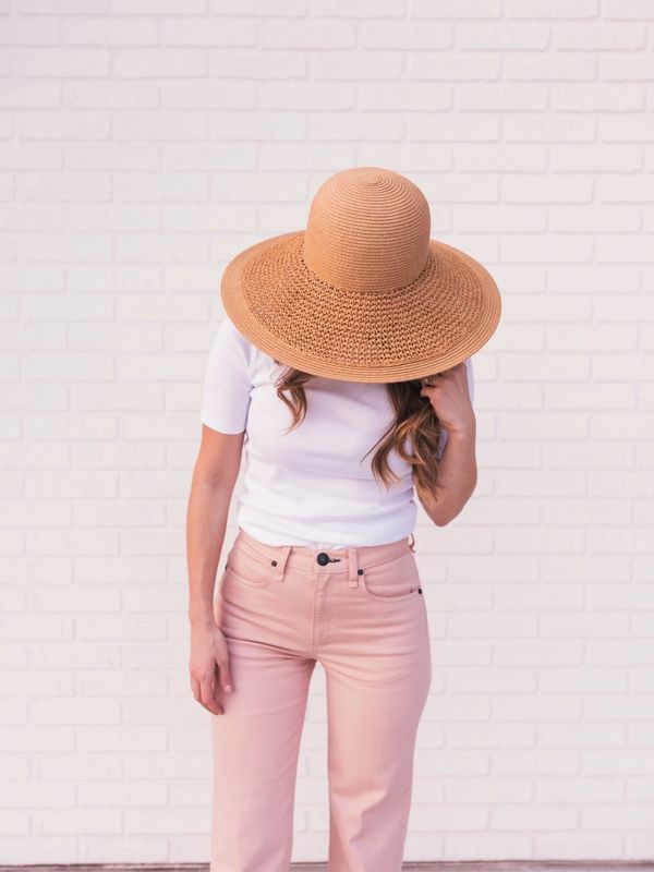 Woman wearing a large straw hat, white shirt, and pink pants against a white brick wall.
