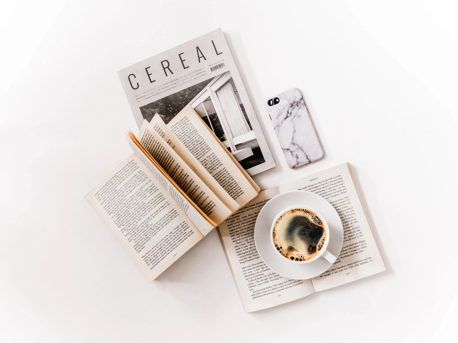 Flat lay of open books, a coffee cup, a magazine, and a marble phone case on a white surface.