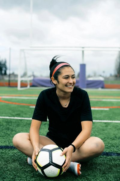 A smiling young woman sits on a soccer field holding a soccer ball.