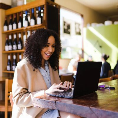 A woman joyfully working on a laptop at a wooden table in a cozy café.