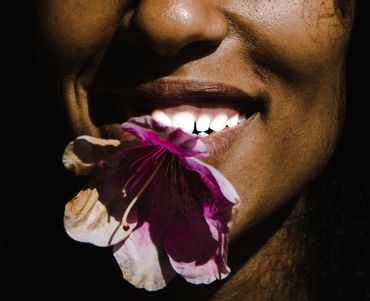 Close-up of a smiling person holding a vibrant purple flower in their mouth.