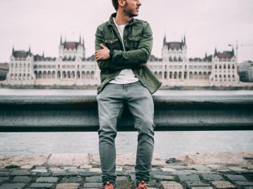 A man stands casually by the river with a grand building in the background.
