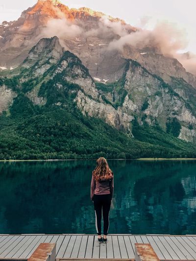 woman standing on a dock facing a mountain landscape and lake