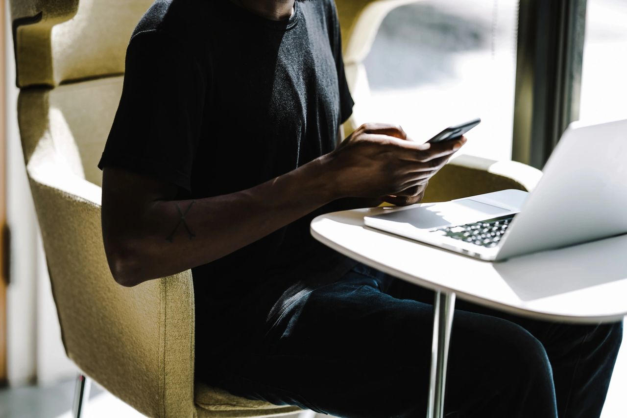 Person sitting in a chair using a smartphone while working on a laptop, illustrating interactive mobile ad engagement and user experience.
