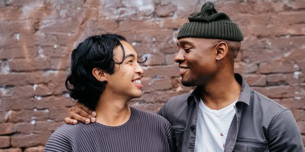 Male presenting couple turned towards each other smiling in front of a brick wall