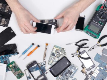 Hands repairing a smartphone surrounded by phone parts and tools.