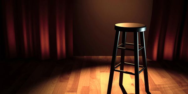 A lone stool under a spotlight on a stage with a red velvet curtain in the background.