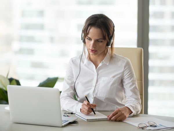 Woman with headset taking notes while working on laptop.