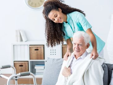 A caregiver gently assists an elderly man seated on a couch.