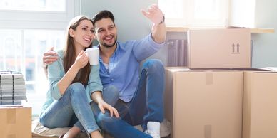 A young man and woman are sitting on the floor in their house, surrounded by packing boxes, smiling.