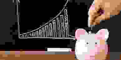 A hand putting a coin into a pink piggy bank in front of a graph drawn in chalk on a blackboard.