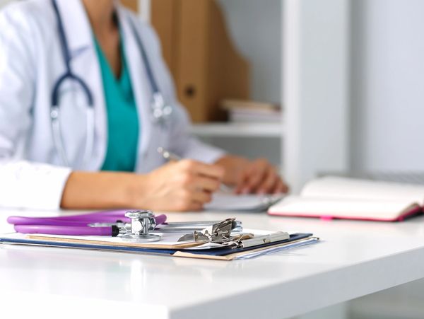 Close-up of a stethoscope and clipboard on a desk with a doctor writing in the background.