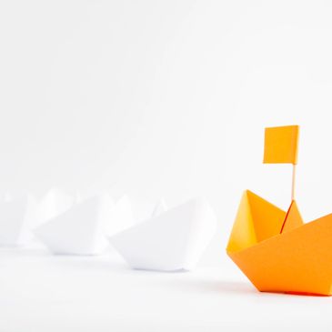 An orange paper boat with a flag leads white paper boats in a row.