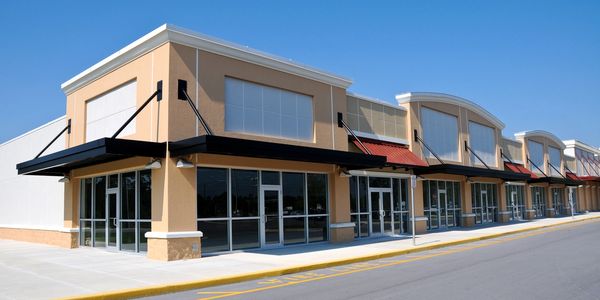 Empty retail storefronts in a strip mall under clear blue sky.
