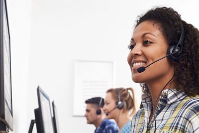 A smiling woman wearing a headset works at a computer in a call center.