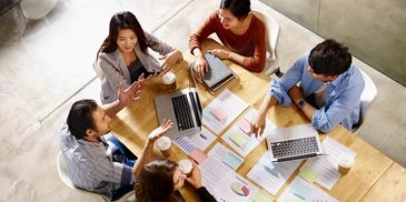 A group of five colleagues collaborating around a table with laptops and documents.