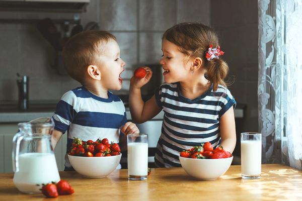 Two children, boy and girl, eating strawberries and cups of milk in a kitchen.