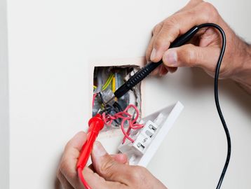 Hands using a multimeter to test electrical wires inside a wall socket.