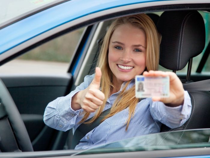 Smiling woman in car showing driver's license and thumbs up.