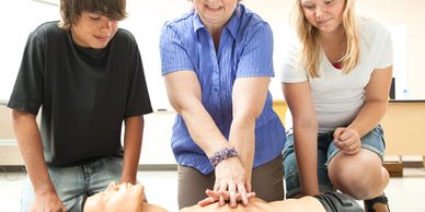 An instructor demonstrates CPR on a mannequin with two students watching closely.