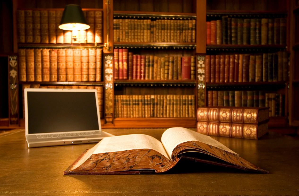 An open book laying on a desk, with a laptop nearby. Background contains a bookcase full of books.