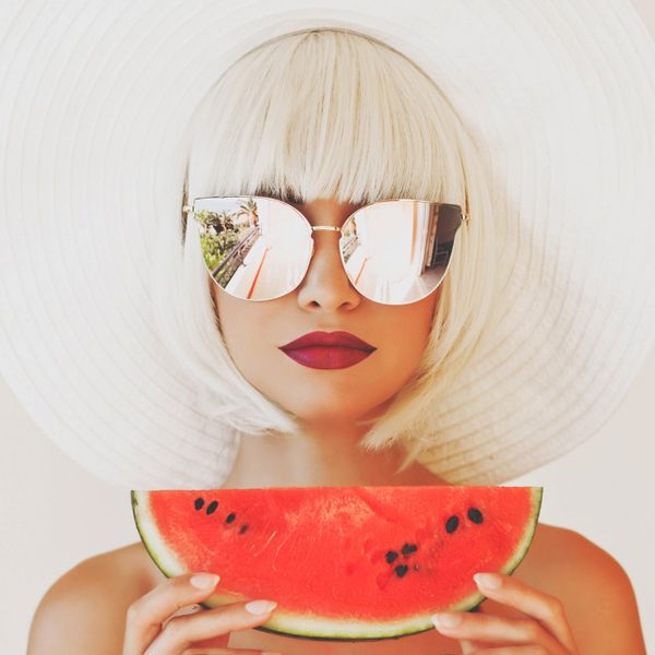 Platinum blonde woman wearing big mirrored sunglasses and red lipstick holding a piece of watermelon