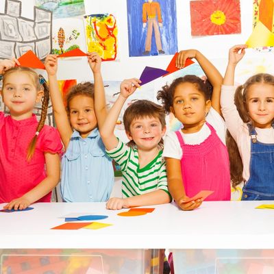 Five children proudly hold up colorful paper crafts in a bright classroom.