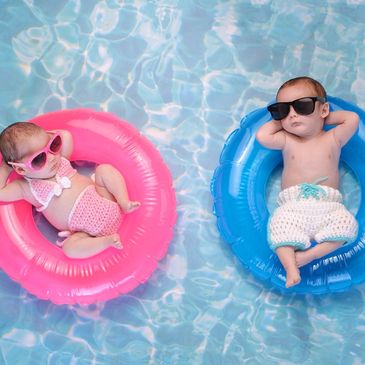 Two babies relaxing on pink and blue floaties in a pool, wearing sunglasses and swimwear.