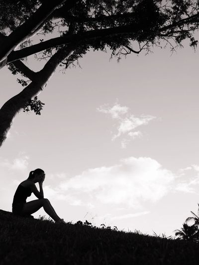 A black and white photo of a woman sitting on a hill, under a tree, in silhouette