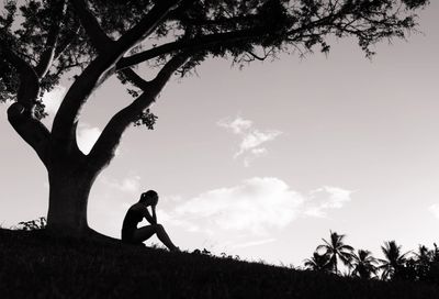 Silhouette of a person sitting under a tree with hands on their face. 