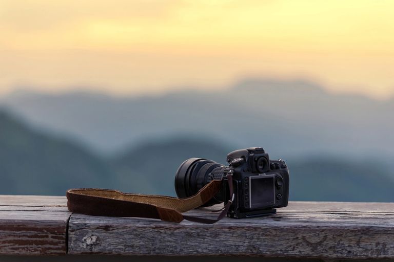 A DSLR stills camera with a leather strap sitting on a wooden ledge with a blurred mountain landscap