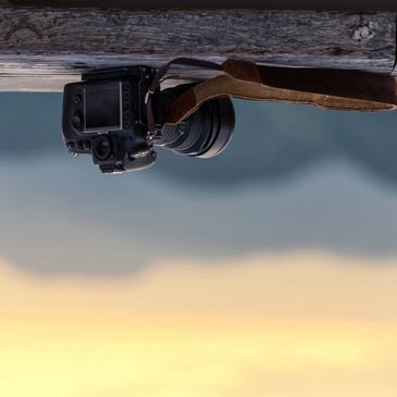 A DSLR camera hanging upside down from a wooden beam against a blurry outdoor background.