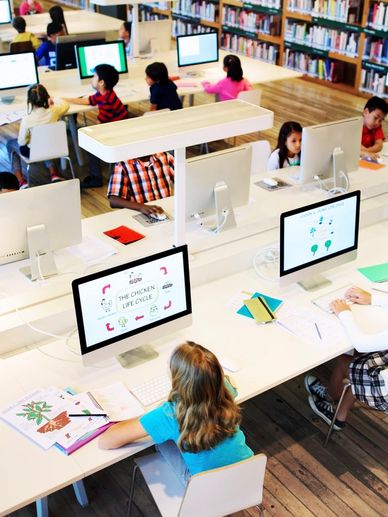 Children learning on computers in a bright, modern classroom with a teacher assisting.