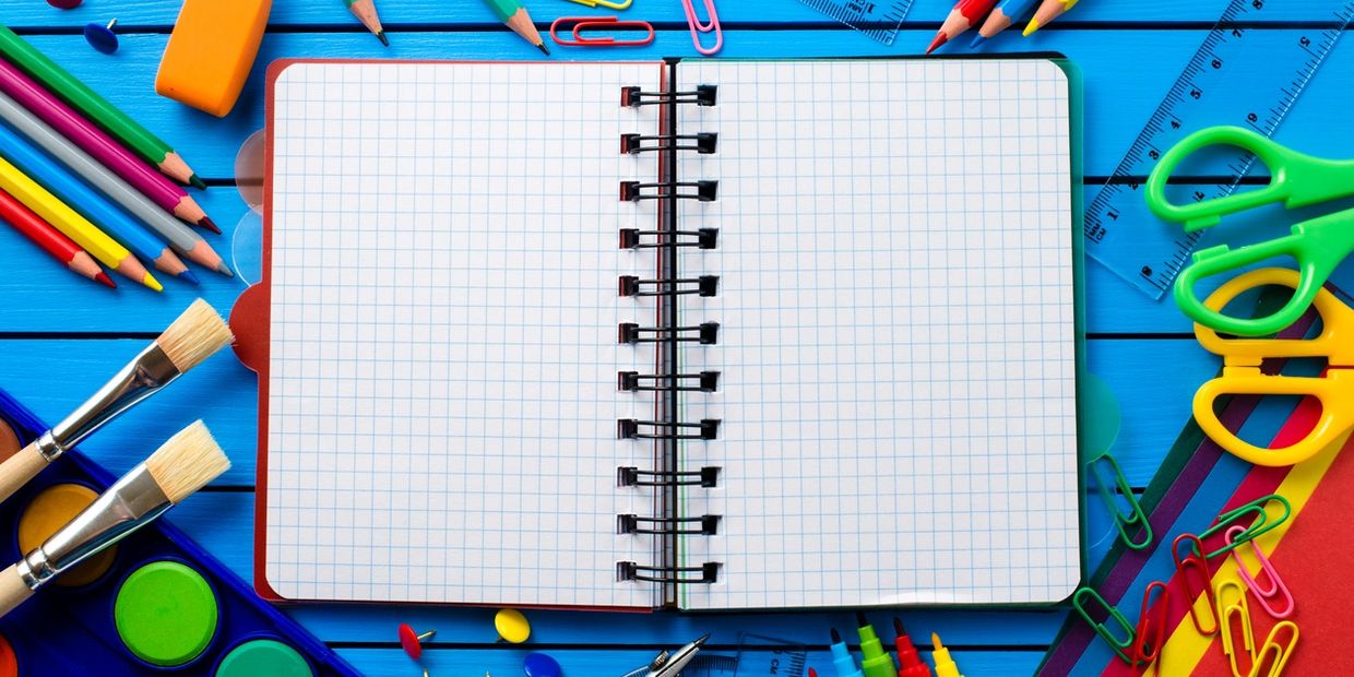 Open grid notebook surrounded by colorful school supplies on blue wooden surface.