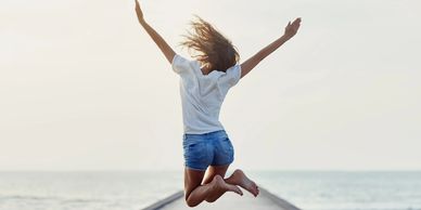 Woman in bare feet, white t-shirt, and shorts jumping for joy on dock surrounded by water