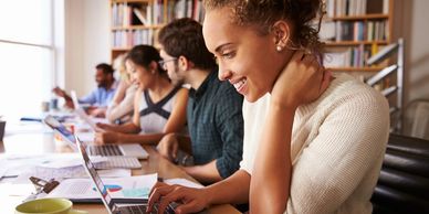 multiple students sitting at large desk at window working on laptops and talking