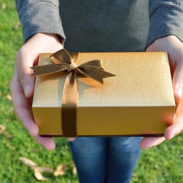 Person holding a gold gift box with a brown ribbon outdoors.