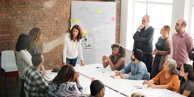 Fourteen adults standing or sitting around a large white table with a whiteboard.