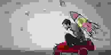 Young man sitting in a red toy car in front of a wall with cartoon space rocket drawn on it.