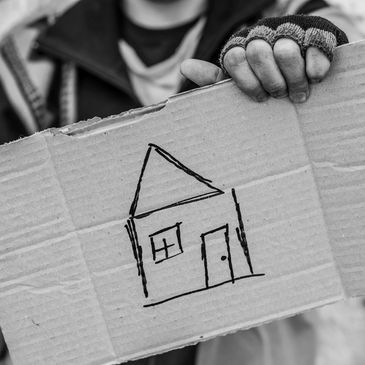 A person in tattered clothes holding a piece of cardboard with a house doodled on it