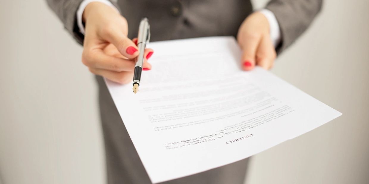 A person handing a pen and contract document to the viewer seemingly to sign